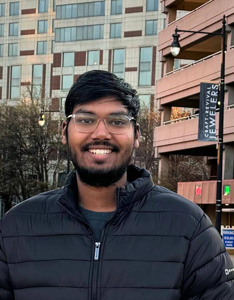 Man with glasses, black and and short black beard smiling at the camera with buildings in the background